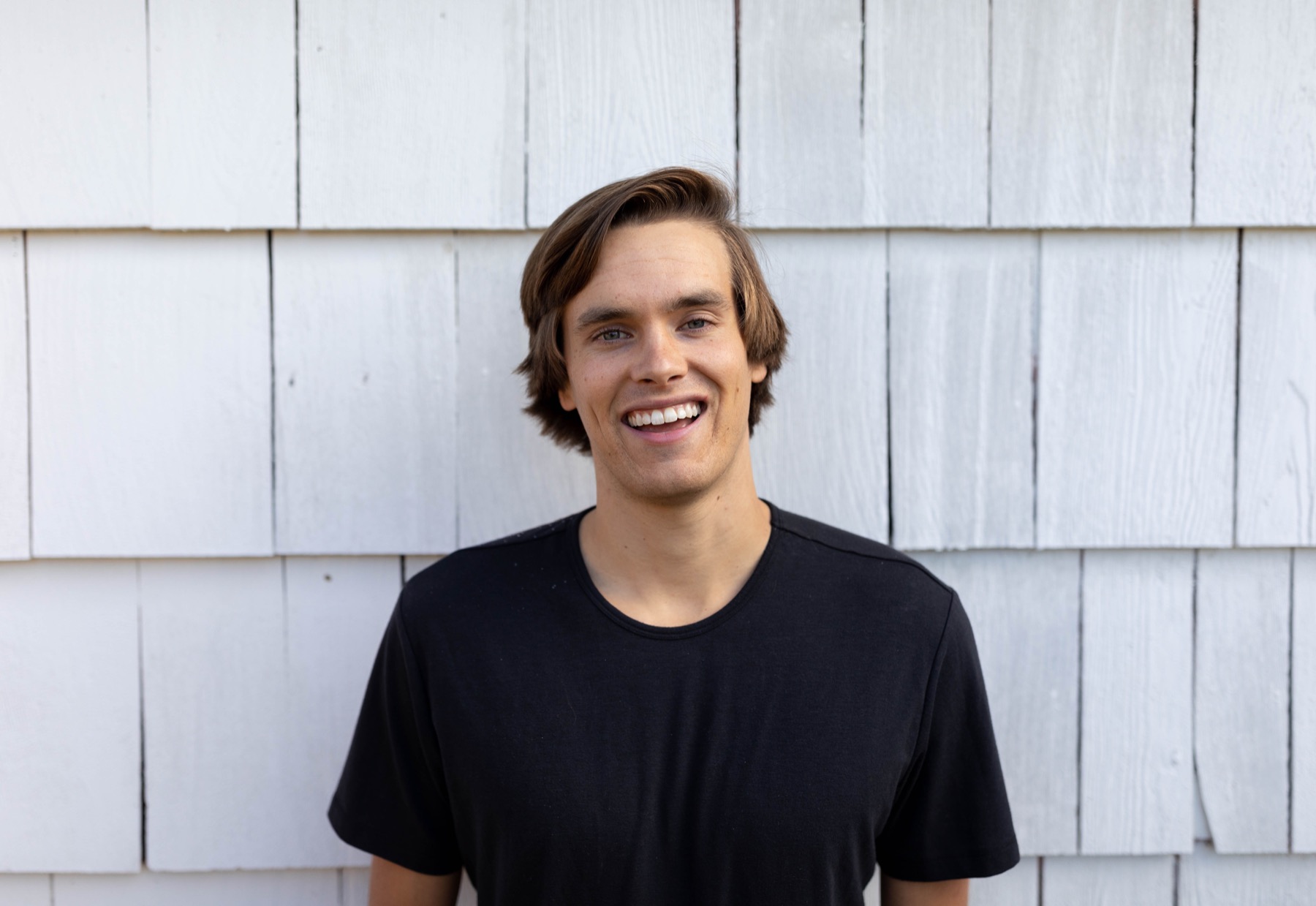 Kory Kirby smiling in a black t-shirt in front of white cedar shingles